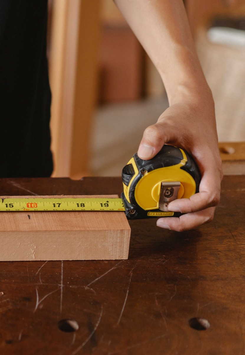 A hand measuring the length of a wooden plank with a tape measure